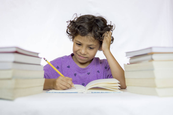 Gelukkige jongen tussen 8 en 10 jaar oud met de uitdrukking van twijfel zittend aan een bureau zijn huiswerk te maken, met leerboeken op zijn tafel en met een witte achtergrond - Foto, afbeelding