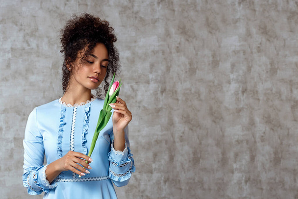 Young African girl in a blue dress with a Tulip on a gray background. The concept of women's day and spring - Photo, Image