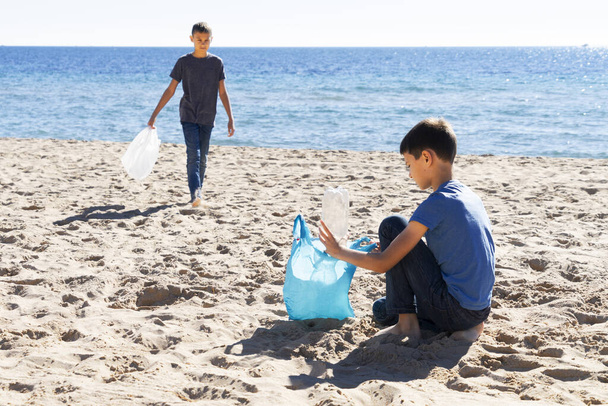 Volunteers cleaning beach from plastic. Boys walking on the beach and picking up plastic bottles trash and putting into plastic bag for recycle - Фото, изображение