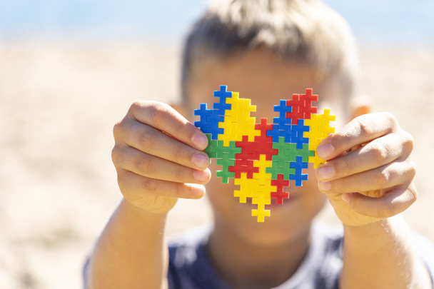 Menino segurando colorido coração de quebra-cabeça na frente de seu rosto. Conceito de Dia Mundial do Autismo
 - Foto, Imagem