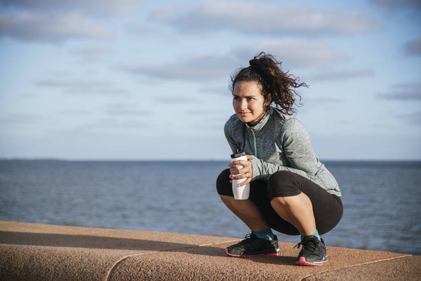 white girl, latin, brunette with curls, drinking water while resting from her training, wearing sportswear, near the sea - Photo, Image