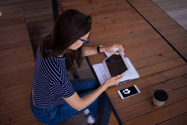 A serious brunette girl, freelancer, with large wrist watch, works at a wooden table on tablet, with gadgets and papers. - Foto, Bild