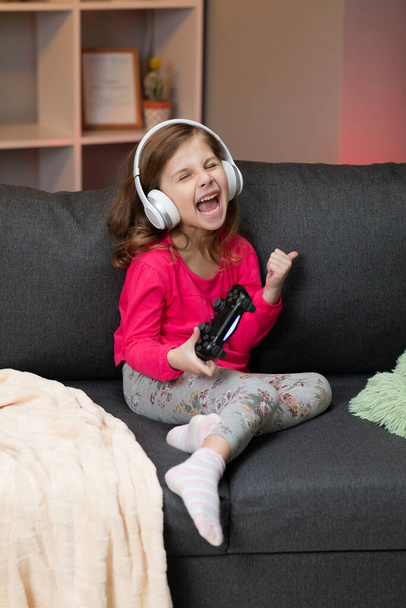 Little girl sitting on sofa playing a video in living room at home. Excited gamer girl hand holding joystick playing console game using a wireless controller. Having fun,enjoy happy expression - Foto, imagen