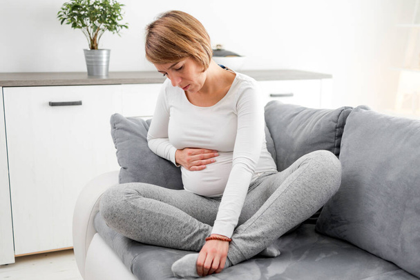 Pregnant tired exhausted woman with stomach, leg and pain issues at home on a couch, being sick. - 写真・画像