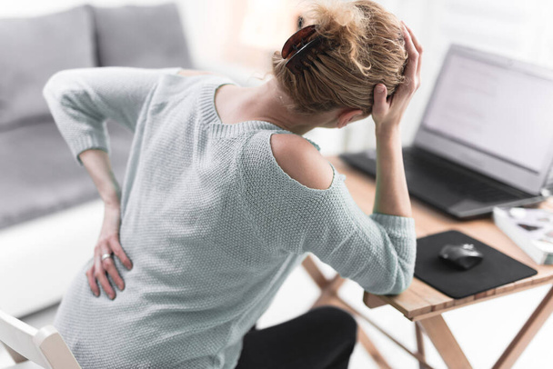 Vrouw die aan een laptop werkt en hoofdpijn en rugpijn heeft, heup, rugpijn. - Foto, afbeelding