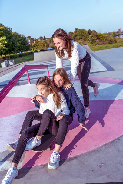 3 girls schoolgirls ride skateboard, rest after school institute college weekend. Casual wear sweater jeans. Summer city, sports ground. Emotions happiness, fun, joy, laughter, smile, entertainment. - 写真・画像