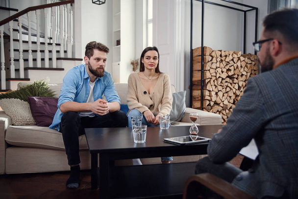 Close up of male psychologist sits at chair and listens depressed young couple at stylish cozy office. Selective focus. - Foto, imagen