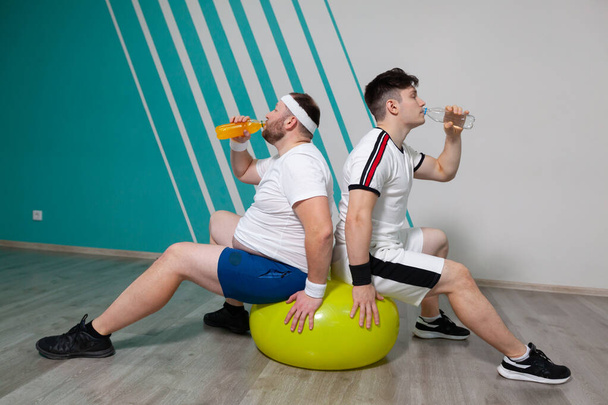 Fat man and his personal trainer are sitting on fitness ball and drinking water after hard training in fitness group classes. The overweight man is extremely tired - Photo, image