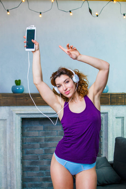 Cheerful young woman in a purple top and blue pants with headphones on is dancing to the music at home - Photo, image