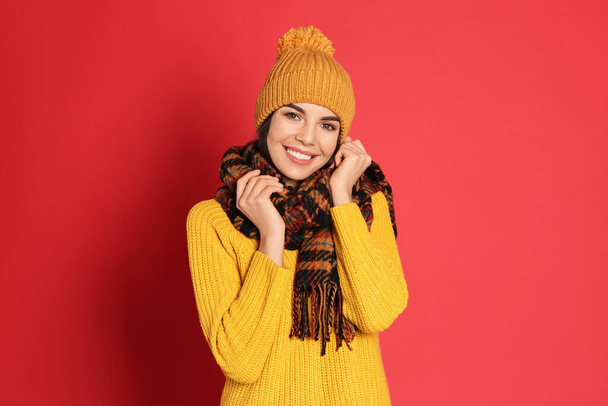Νεαρή γυναίκα που φοράει ζεστό πουλόβερ, κασκόλ και καπέλο σε κόκκινο φόντο. Χειμερινή περίοδος - Φωτογραφία, εικόνα