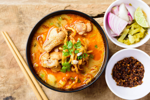 Традиционная северная тайская еда (Khao Soi), острый суп с лапшой карри с кокосовым молоком и курицей, поедающая хрустящую яичную лапшу, маринованную горчичную зелень, лук-шалот, лайм, листья кориандра и молотый чили, жареный в масле. Вид сверху
 - Фото, изображение
