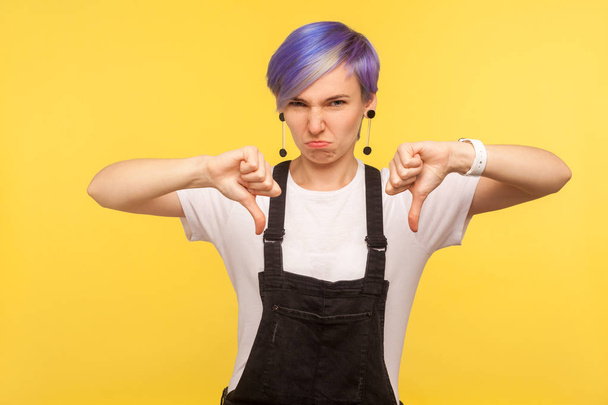 Dislike! Portrait of dissatisfied hipster girl with violet short hair in denim overalls frowning with displeasure and showing thumbs down, negative disapproval sign. yellow background, studio shot - Zdjęcie, obraz