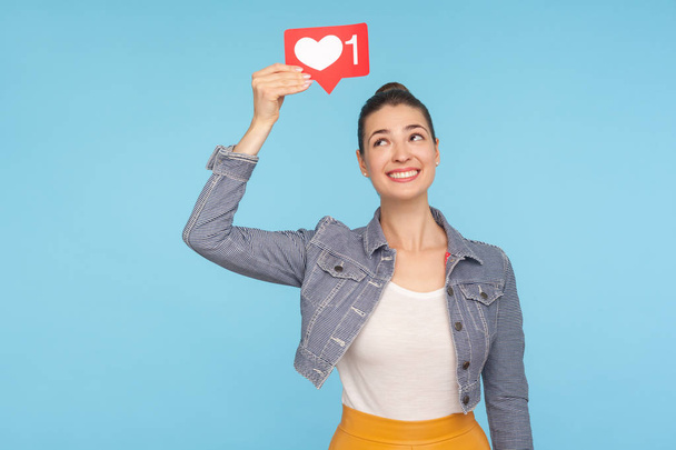 Use network Like button! Portrait of happy fashionably dressed woman with hair bun holding social media heart icon over head and looking at it, recommending to follow, love content. studio shot - Foto, immagini