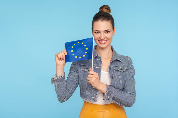Symbol of Europe, EU association. Beautiful friendly fashionably dressed woman with hair bun smiling broadly and holding flag of European Union, community. studio shot isolated on blue background - Foto, immagini