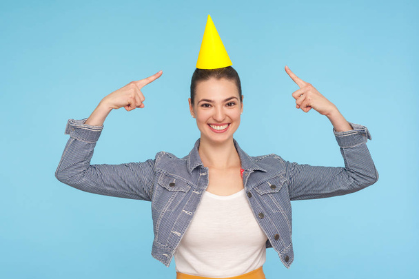 Fascinating cute happy woman in denim jacket pointing at funny party cone on her head and smiling at camera, feeling like happy birthday girl, winner. indoor studio shot isolated on blue background - Photo, image