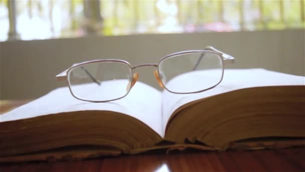 Egy nyitott könyv és egy szemüveg vagy szemüveg a fa asztalon, egy meleg napos reggelen, oldalnézetből közelről. Vasárnap ünnepi koncepció. Elszigetelt háttér szelektív fókuszban sekély mélységű mezővel. - Felvétel, videó