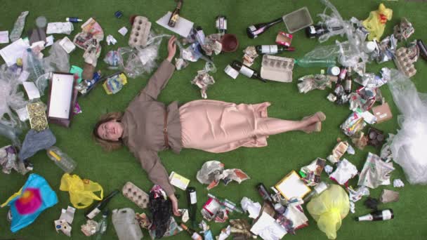 Holka mezi odpadky. Krásná dívka leží na trávě mezi různými odpadními produkty: skleněné nádoby, papírový odpad, obaly, plast, jednorázové nádobí, plechovky, láhve, krabice. - Záběry, video