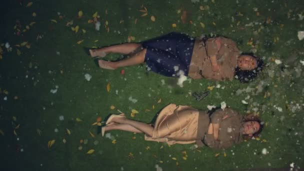 Two girls on the grass. Slow motion 2x. Top view.Asian and caucasian girls lie on the grass among the yellow leaves. Between them is a gun. Evening. Snowing. - Materiaali, video