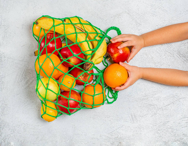 Children's hands put fruit in a string bag with mixed fruits: bananaite, lemons, apples, oranges. Zero waste concept. Gray background, copy space, closeup. View from above. - Foto, afbeelding