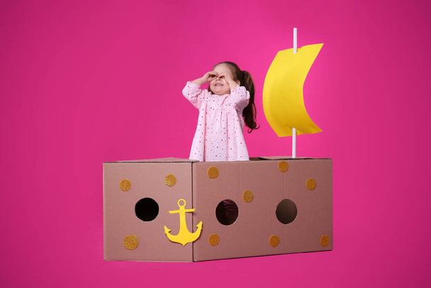 Little child playing with ship made of cardboard box on pink background - Foto, imagen