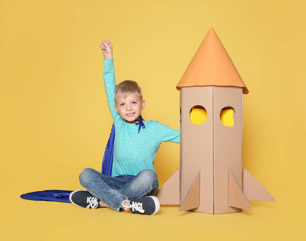 Little child in cape playing with rocket made of cardboard box on yellow background - Fotó, kép