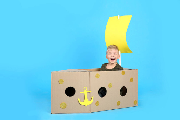 Little child playing with ship made of cardboard box on light blue background - Photo, Image