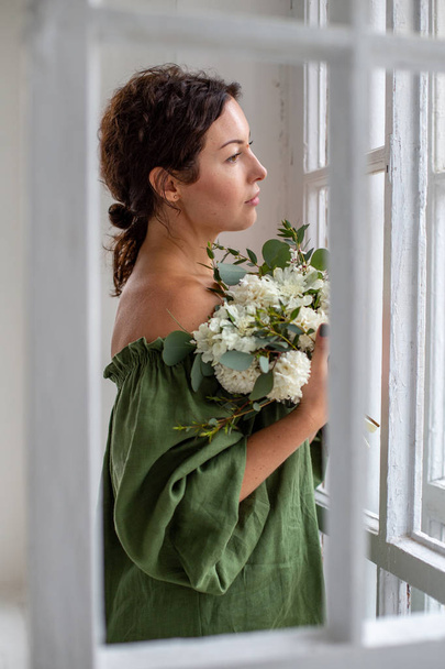 κορίτσι σε ένα πράσινο φόρεμα με ένα μπουκέτο υάκινθους στα χέρια της στέκεται δίπλα στο παράθυρο και κοιτάζει δυστυχώς στην απόσταση - Φωτογραφία, εικόνα