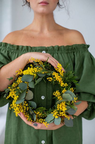 Foto einer schönen Frau in einem grünen Kleid mit nackten Schultern und einem Kranz aus Mimosen in ihren Händen - Foto, Bild