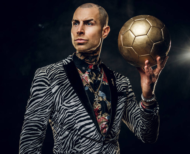 Смілива татуйована чоловіча модель позує для фотосесії в темній студії і тримає золотий футбольний м'яч, дивлячись вбік
 - Фото, зображення