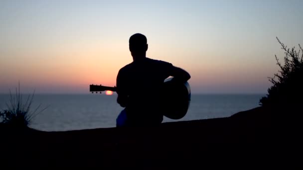 Sylwetka młodego mężczyzny piszącego piosenki na świeżym powietrzu grającego na gitarze akustycznej o zachodzie słońca - Materiał filmowy, wideo