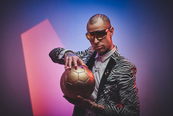 Merész tetovált férfi modell pózol fotózáshoz egy sötét stúdióban, és egy arany focilabdát tart, miközben mosolyog - Fotó, kép
