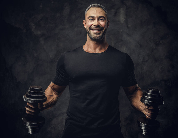 Serious, adult, fit muscular caucasian man coach posing for a photoshoot in a dark studio, wearing sportswear, showing his muscles with dumbbells - Zdjęcie, obraz