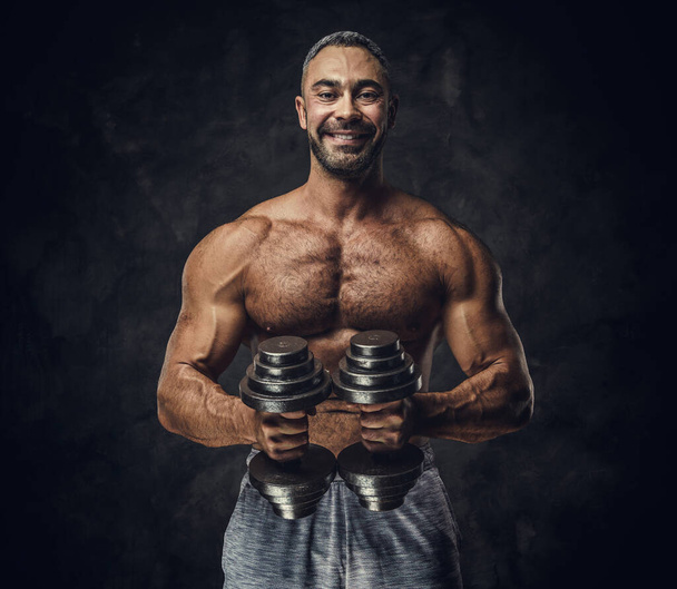 Charming, adult, fit muscular caucasian man coach posing for a photoshoot in a dark studio, wearing sportswear, showing his muscles with dumbbells - Fotoğraf, Görsel
