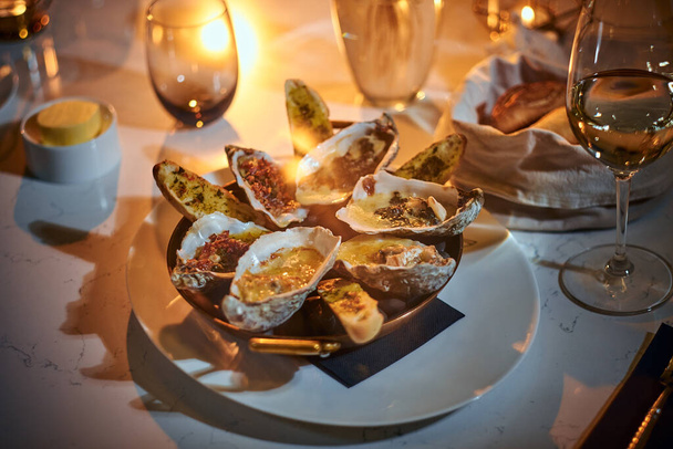 Haute cuisine dinner bowl of oysters and bread loafs with sauce served on a table next to candle, a glass of wine, and a wicker basket of bread in a restaurant - 写真・画像