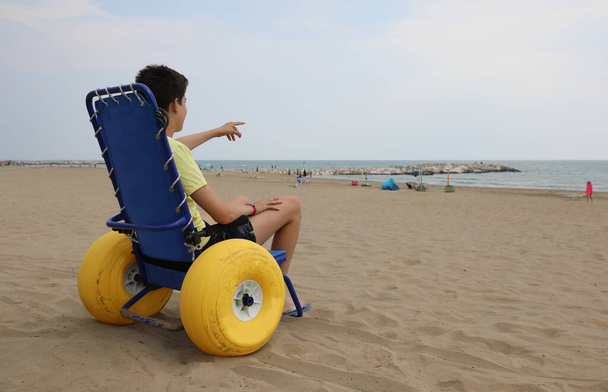 夏に海に行くために大きなインフレータブルホイールを持つ特別な車椅子の少年 - 写真・画像