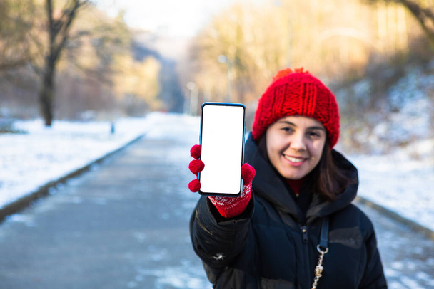 young smiling woman in winter outfit holding phone with white blank empty screen copy space - Photo, Image