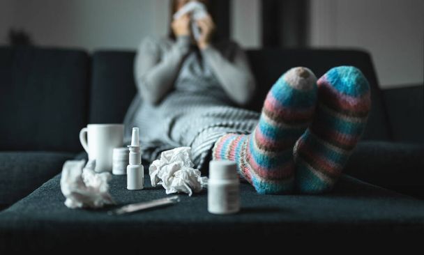 Frau niest und pustet Nase mit Gewebe und Taschentuch. Kranke und Kranke mit Grippe, Erkältungsmedikamenten und Wollsocken. Fieber, Viren oder Infektionen. Im Winter zu Hause auf der Couch sitzen. - Foto, Bild