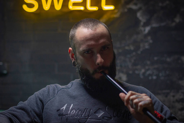 γενειοφόρος άντρας που καπνίζει ναργιλέ σε σκοτεινό δωμάτιο. τρόπος ζωής hookah και την έννοια του καπνίσματος. πορτρέτο ενός ναύτη με ένα hookah από κοντά - Φωτογραφία, εικόνα
