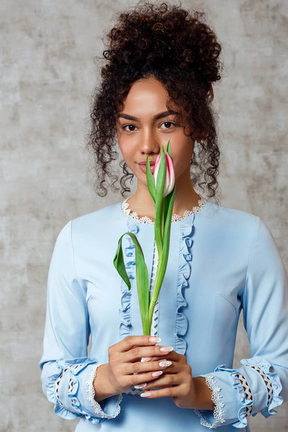Young African girl in a blue dress with a Tulip on a gray background. The concept of women's day and spring - Foto, imagen