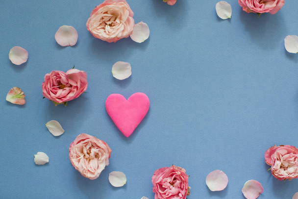 Corazón de color rosa brillante dentro del marco de rosas y pétalos sobre un fondo azul. Vista superior
 - Foto, imagen