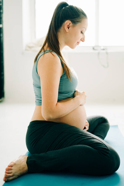 gyönyörű terhes lány sportruházatban ül egy jóga matracon és simogatja a hasát - Fotó, kép