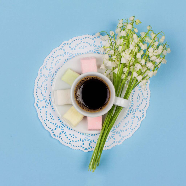 white cup of coffee latte on a saucer, marshmallow, a bouquet of lily of the valley flowers on blue background, top view flat lay - Foto, afbeelding