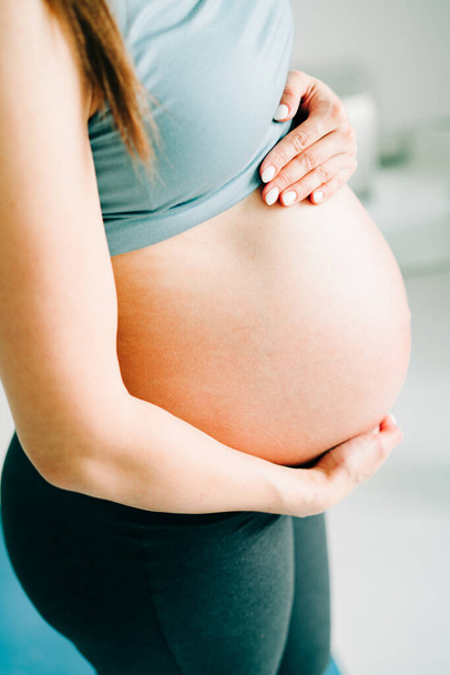 mladá těhotná dívka ve sportovním oblečení stojí na józe Mat, usmívá se a drží se za břicho - Fotografie, Obrázek