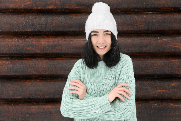 Young woman wearing warm sweater and hat near wooden wall. Winter season - Valokuva, kuva