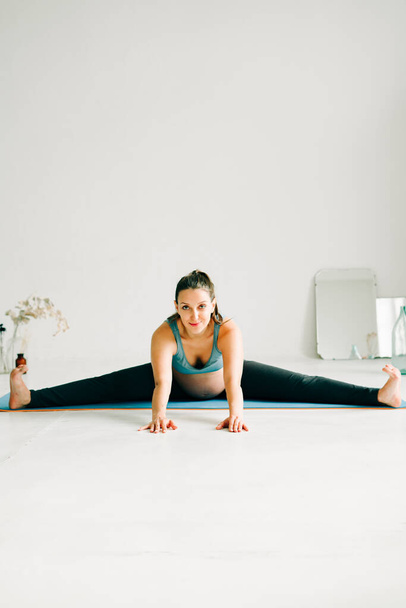 eine junge schwangere Frau in Sportkleidung macht Yoga und macht Übungen auf einem Teppich sitzend - Foto, Bild