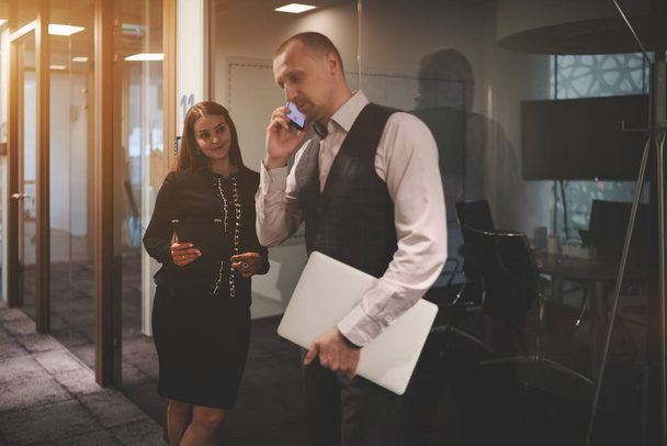 Two business partners are waiting for the beginning of a meeting in an office corridor near the entrance to a board room; selective focus on the woman entrepreneur, her colleague is phoning near her - 写真・画像