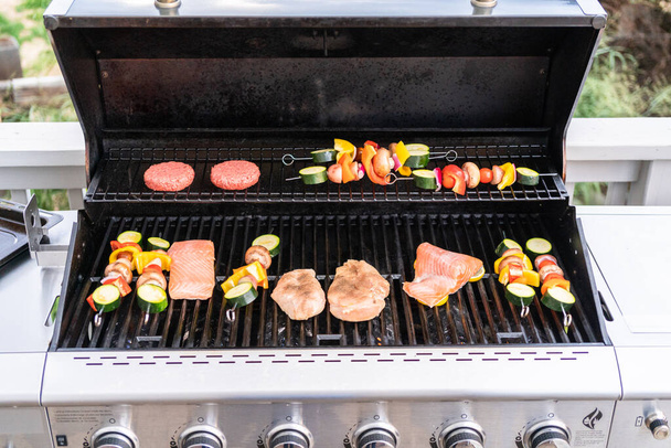 Grilling an Atlantic salmon, chicken breast, vegetable skewers, and vegetarian burgers on an outdoor grill. - Photo, image