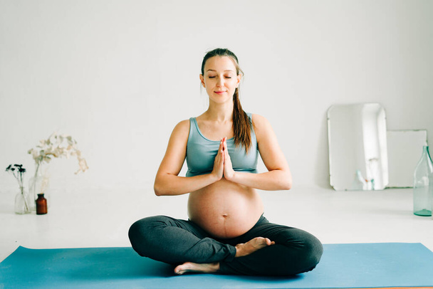 a young beautiful pregnant girl in sportswear performs a meditation practice sitting on a rug - Foto, Imagem