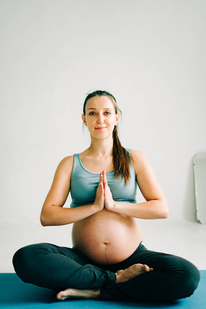 a young beautiful pregnant girl in sportswear performs a meditation practice sitting on a rug - Photo, Image