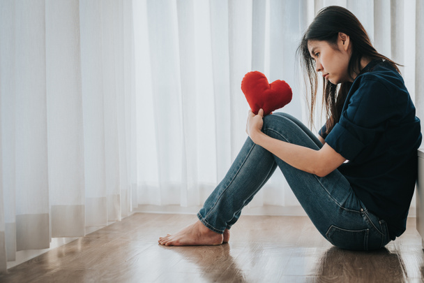 Triste mujer solitaria y deprimida sentada junto a la cama sosteniendo almohada en forma de corazón rojo en su mano
 - Foto, Imagen